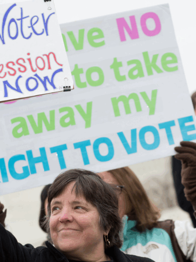A white woman holding a sign that says "End Voter Suppression Now"