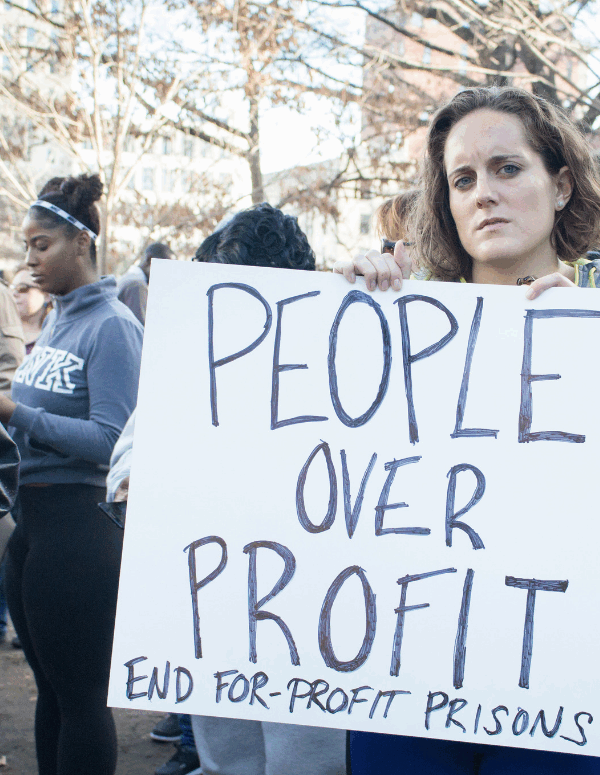 a white woman holding a sign that says "People Over Profit: End For-Profit Prisons)