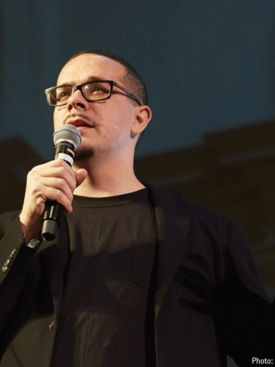 picture of Shaun King speaking at a public event. He dressed in black against a black background.