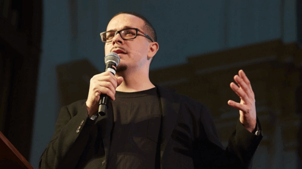 picture of Shaun King speaking at a public event. He dressed in black against a black background.