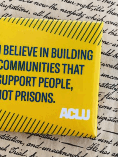 a square yellow button with green text that says "I believe in building communities that support people, not prisons" against a background of cursive writing on parchment paper.