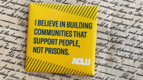 a square yellow button with green text that says "I believe in building communities that support people, not prisons" against a background of cursive writing on parchment paper.