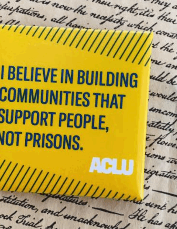 a square yellow button with green text that says "I believe in building communities that support people, not prisons" against a background of cursive writing on parchment paper.
