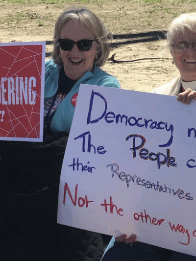 two protesters holding signs that say "End gerrymandering now" and "Democracy means the people choose their reps, not the other way around"