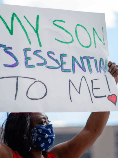 a black woman wearing mask and holding a sign that says "my son is essential to me"