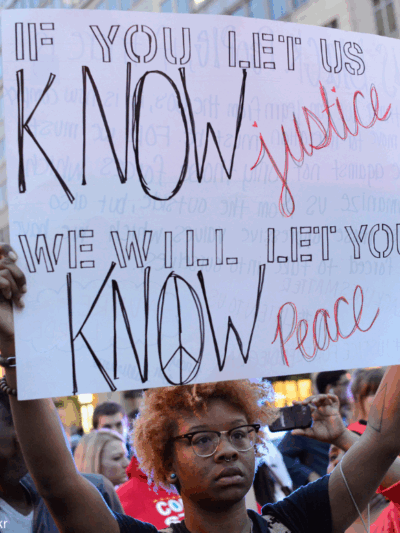 a Black protester holding a sign that says "if you let us know justice, we will let you know peace"