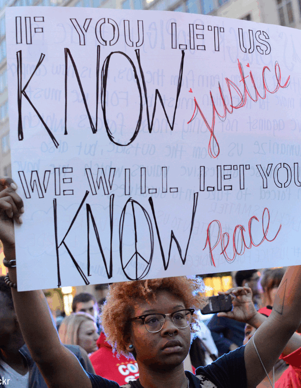 a Black protester holding a sign that says "if you let us know justice, we will let you know peace"