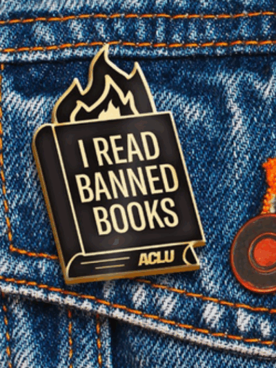 an ACLU branded "I read banned books" pin with fire coming out of a book, against a blue jeans jacket pocket.