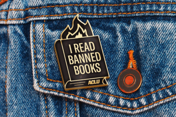 an ACLU branded "I read banned books" pin with fire coming out of a book, against a blue jeans jacket pocket.