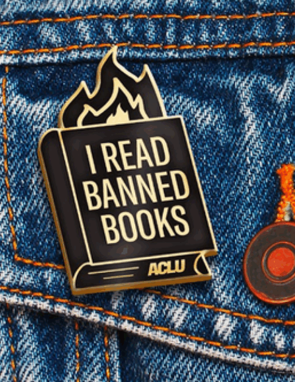 an ACLU branded "I read banned books" pin with fire coming out of a book, against a blue jeans jacket pocket.