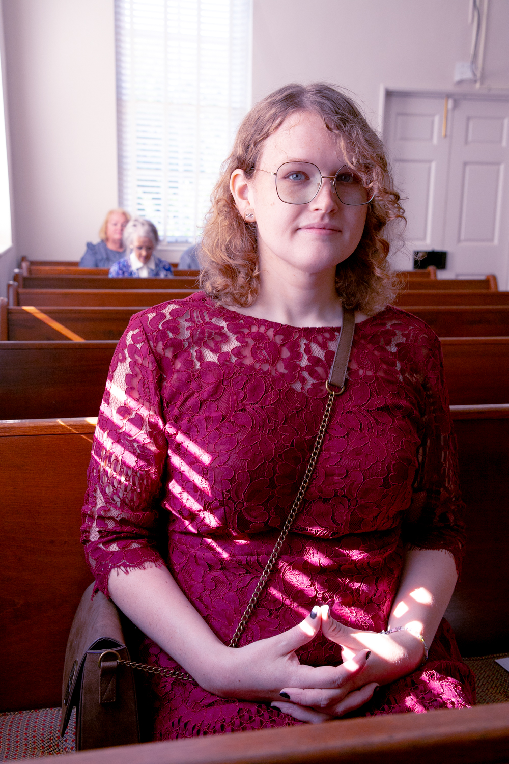 Ellenor ZInksi, wearing a burgundy dress, smiles warmly to the camera with the sunlight pouring over from the window as she sits in church ahead of service.