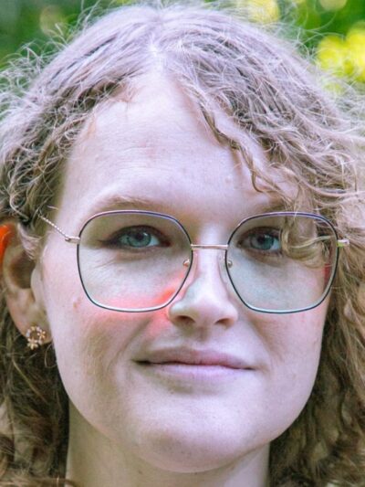 Ellenor, wearing glasses with curly golden hair, smiles confidently while being outside