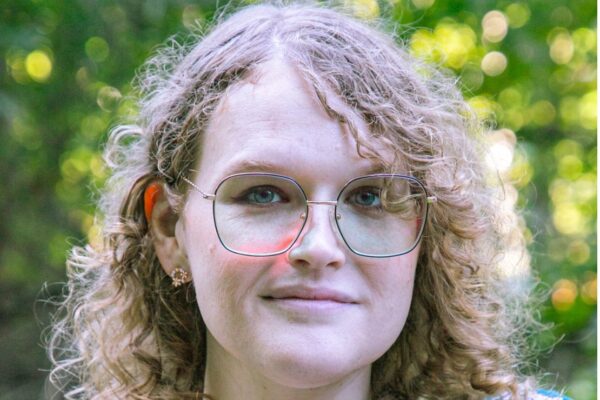 Ellenor, wearing glasses with curly golden hair, smiles confidently while being outside