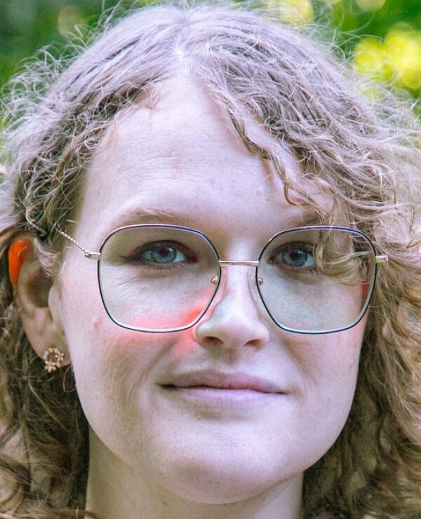 Ellenor, wearing glasses with curly golden hair, smiles confidently while being outside