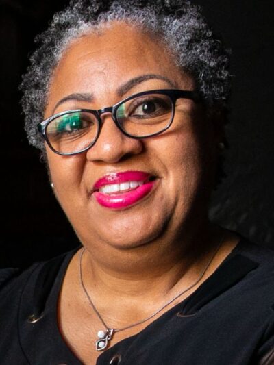 A Black woman with short curly hair and black glasses smiles towards a camera. She is seen holding cards from a loved one.
