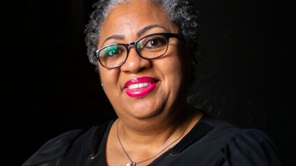 A Black woman with short curly hair and black glasses smiles towards a camera. She is seen holding cards from a loved one.