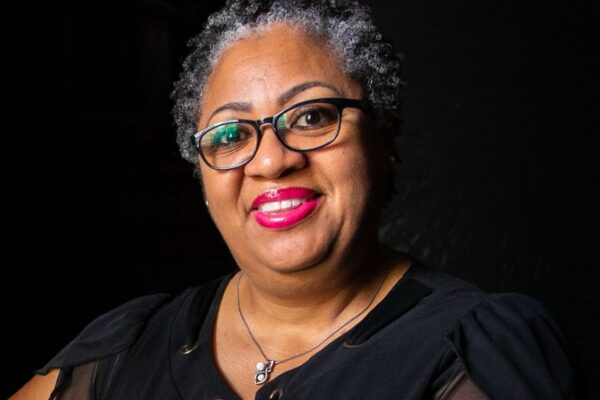 A Black woman with short curly hair and black glasses smiles towards a camera. She is seen holding cards from a loved one.