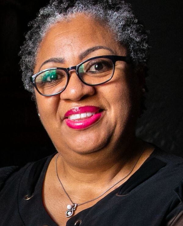 A Black woman with short curly hair and black glasses smiles towards a camera. She is seen holding cards from a loved one.