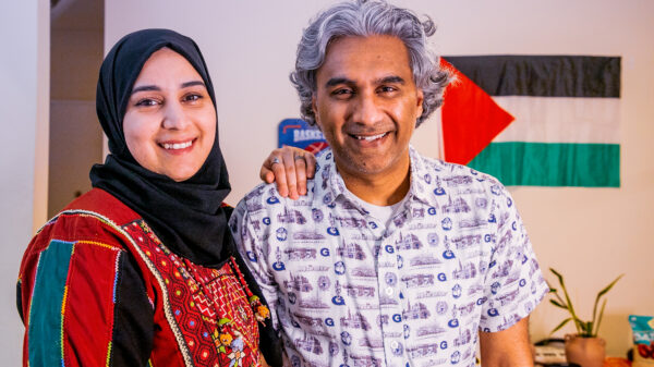 Badar stands next to his wife and smiles with the Palestinian flag in the background