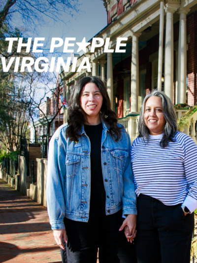 Two people on a cozy sunny sidewalk look towards a camera smiling.
