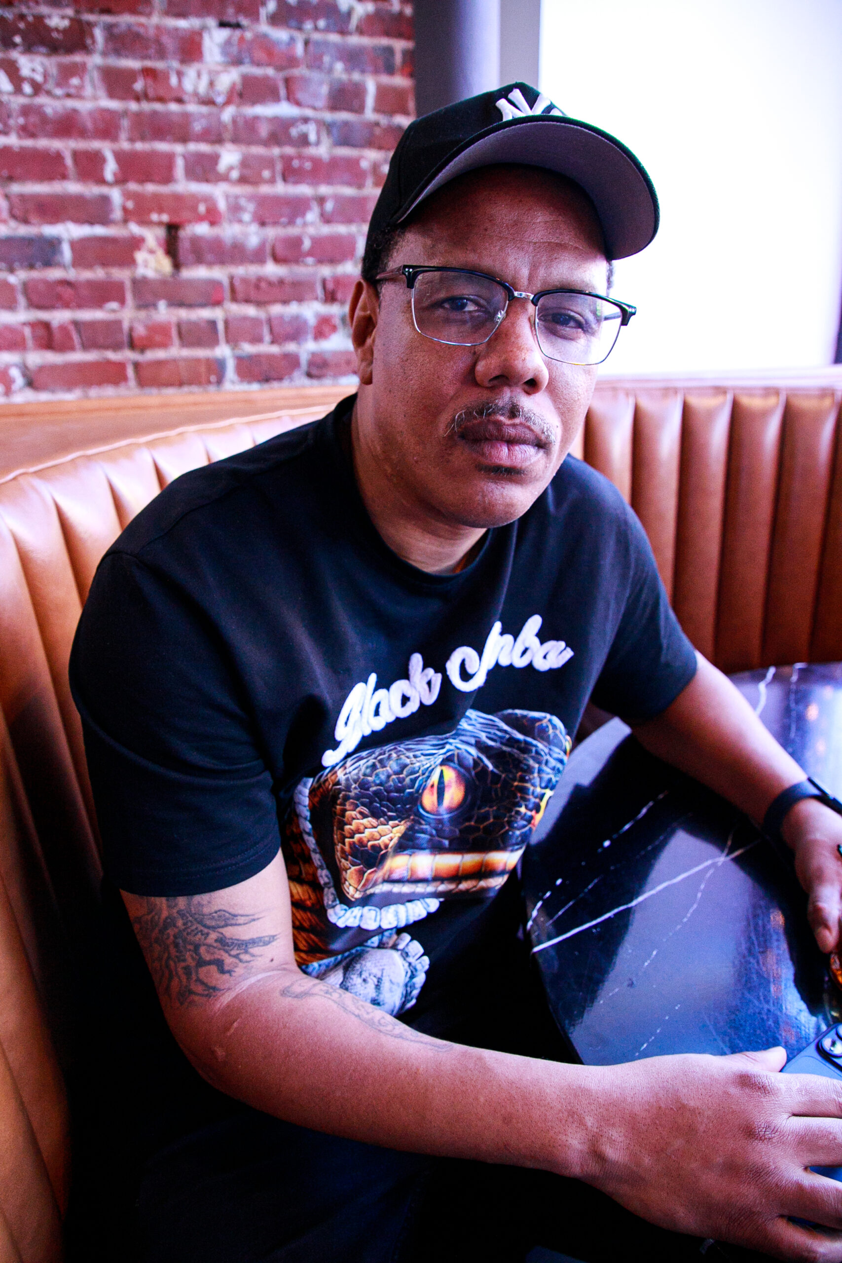 Kenneth Hunter wears a black t-shirt and a black cap sitting at the table at a bar.