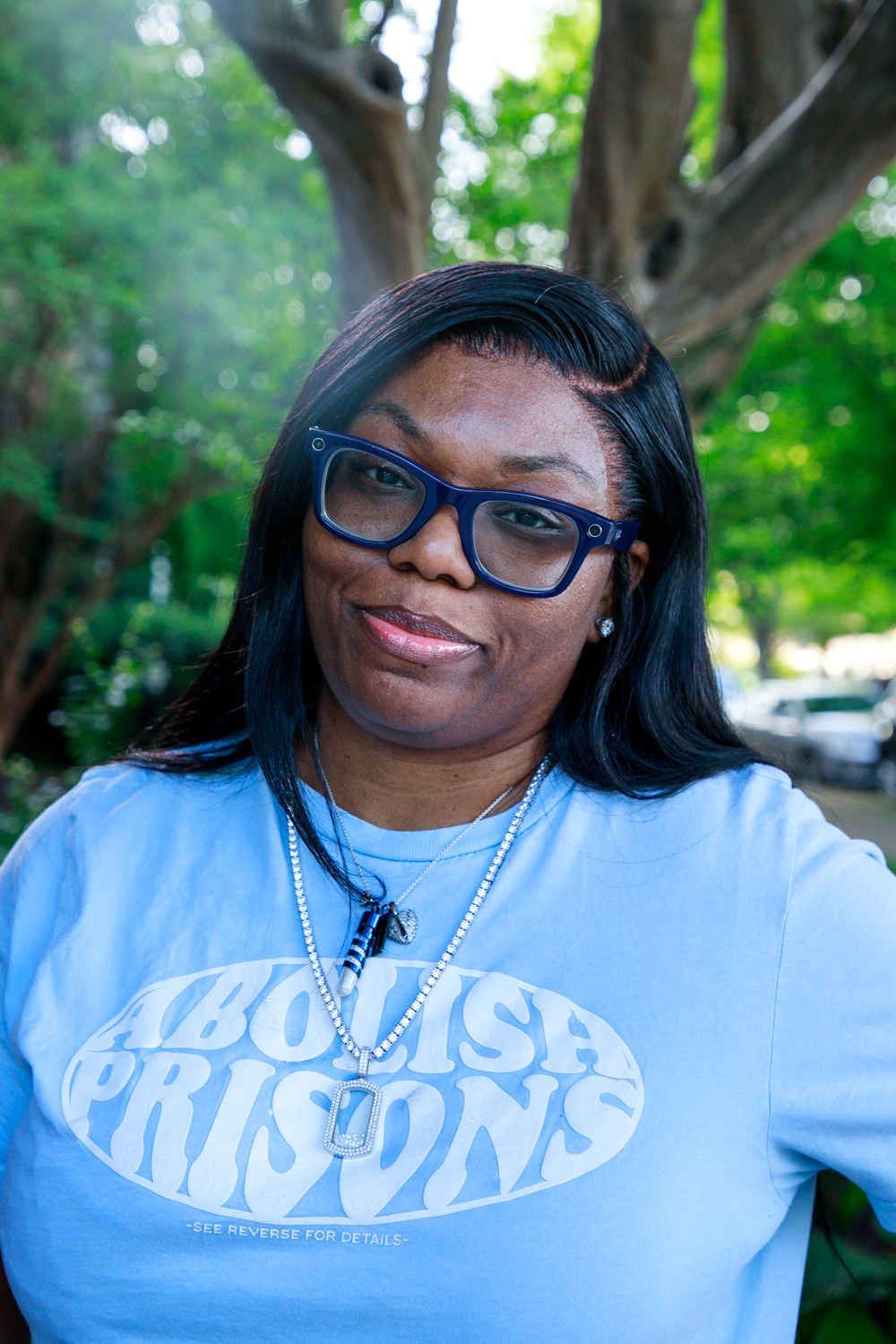 Sheba, a Black woman with long straight hair and black-rimmed glasses, wears an "Abolish Prisons" t-shirt.