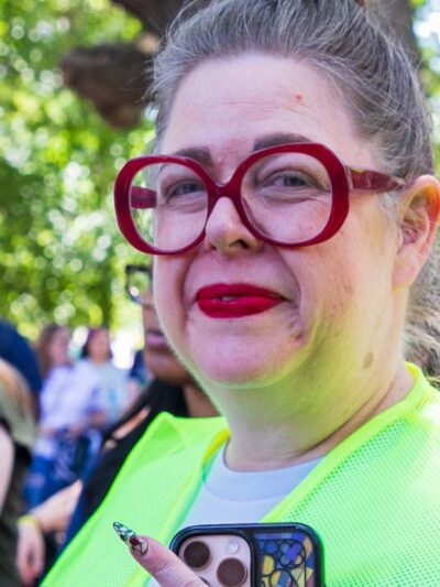 Heather, wearing bright red lipstick and matching red glasses, smiles brightly at the camera