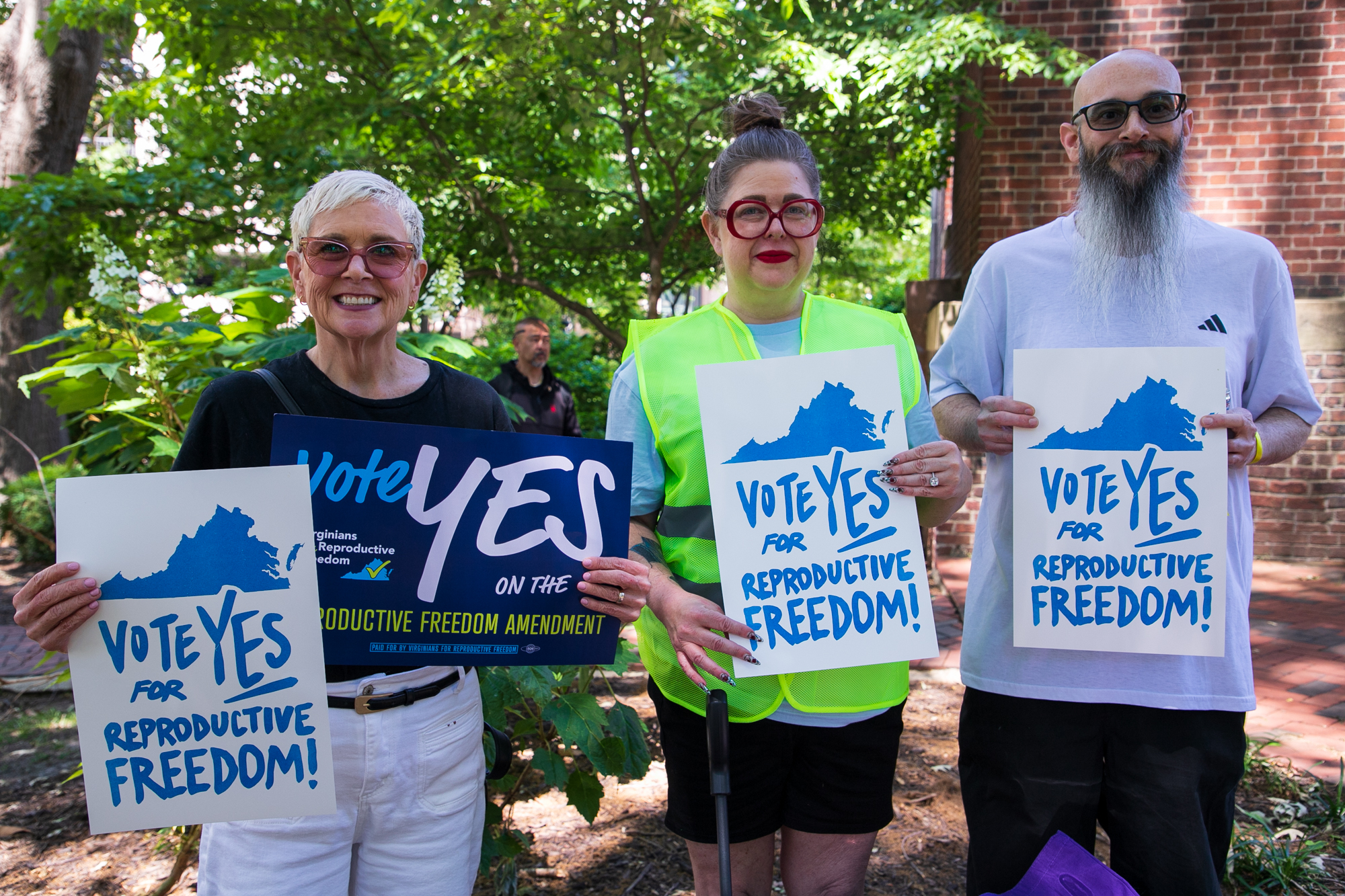 Heather, the ACLU-VA Executive Director Mary Bauer, and her husband hold signs that say "Vote YES for reproductive freedom" at a rally at the Bell Tower on Capitol Grounds