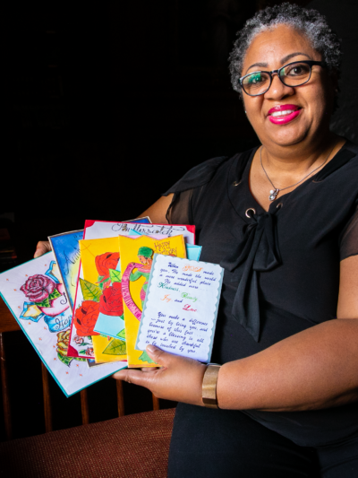 A Black woman with short curly hair and black glasses smiles towards a camera. She is seen holding cards from a loved one.