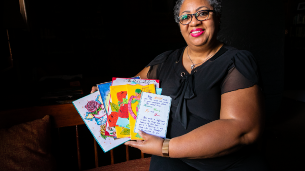 A Black woman with short curly hair and black glasses smiles towards a camera. She is seen holding cards from a loved one.