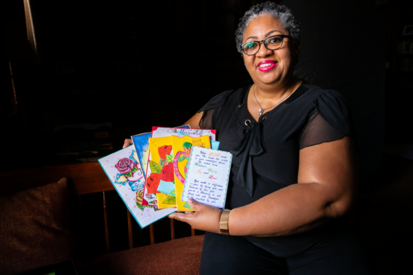 A Black woman with short curly hair and black glasses smiles towards a camera. She is seen holding cards from a loved one.