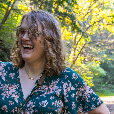 A portrait of a curly haired blonde white woman smiling towards a camera.