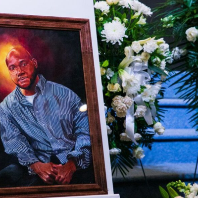 A painting of a black man is set next to a casket and bouquet of flowers.