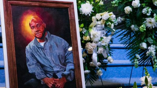 A painting of a black man is set next to a casket and bouquet of flowers.