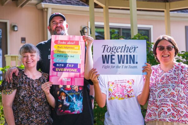 Three adults and two teenagers holding up signs to stay anonymous smiling at the camera