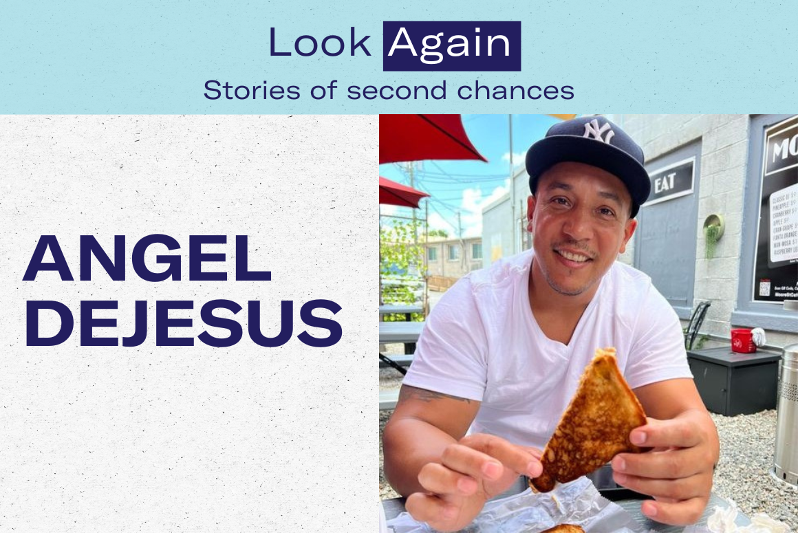 Photo of Angel DeJesus, a Puerto Rican-American man in his 40s, wearing a Yankee baseball cap, white tee, eating his first slice of pizza after prison, smiling brightly to the camera.