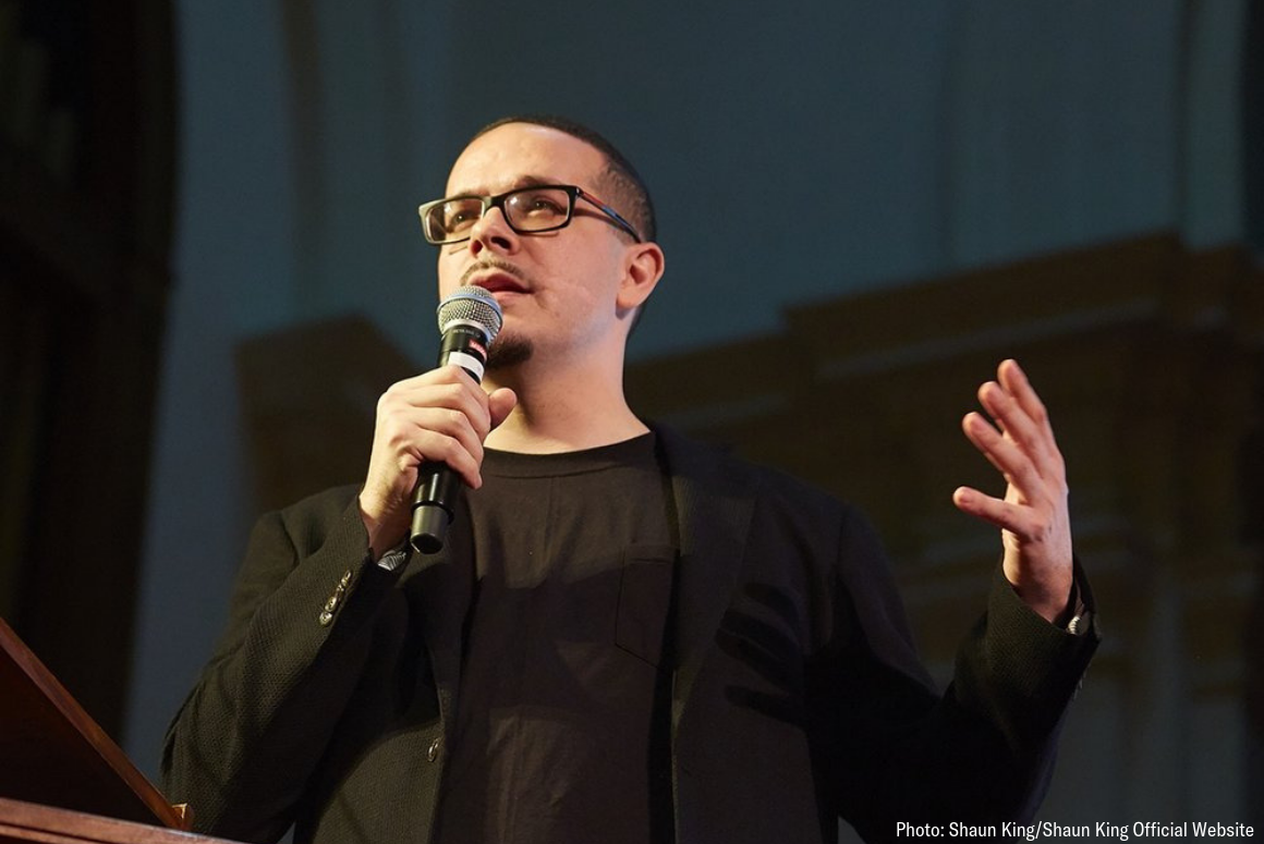 picture of Shaun King speaking at a public event. He dressed in black against a black background.