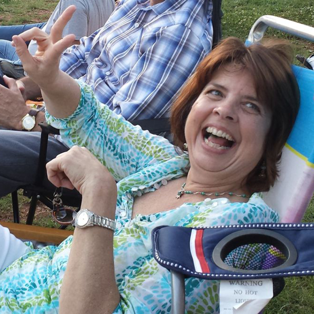 photo of Gay Ellen Plack, a white middle-aged woman, sitting on a lawn chair, laughing heartily.