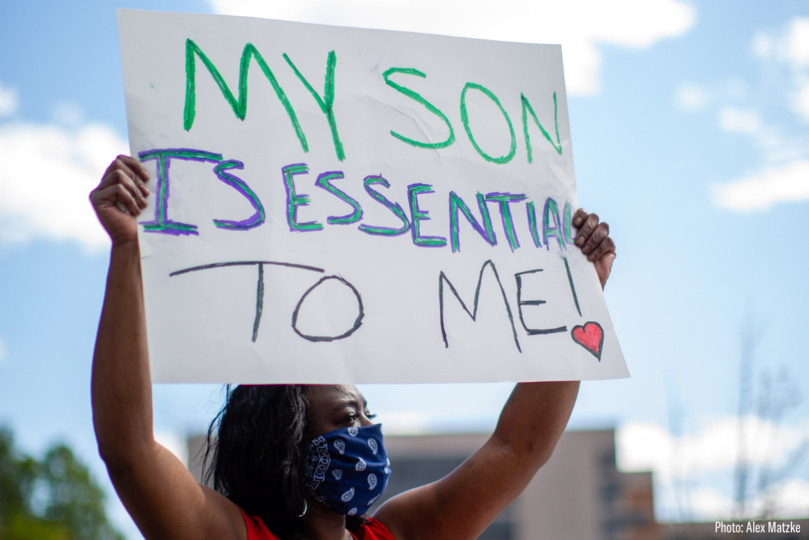 a black woman wearing mask and holding a sign that says "my son is essential to me"