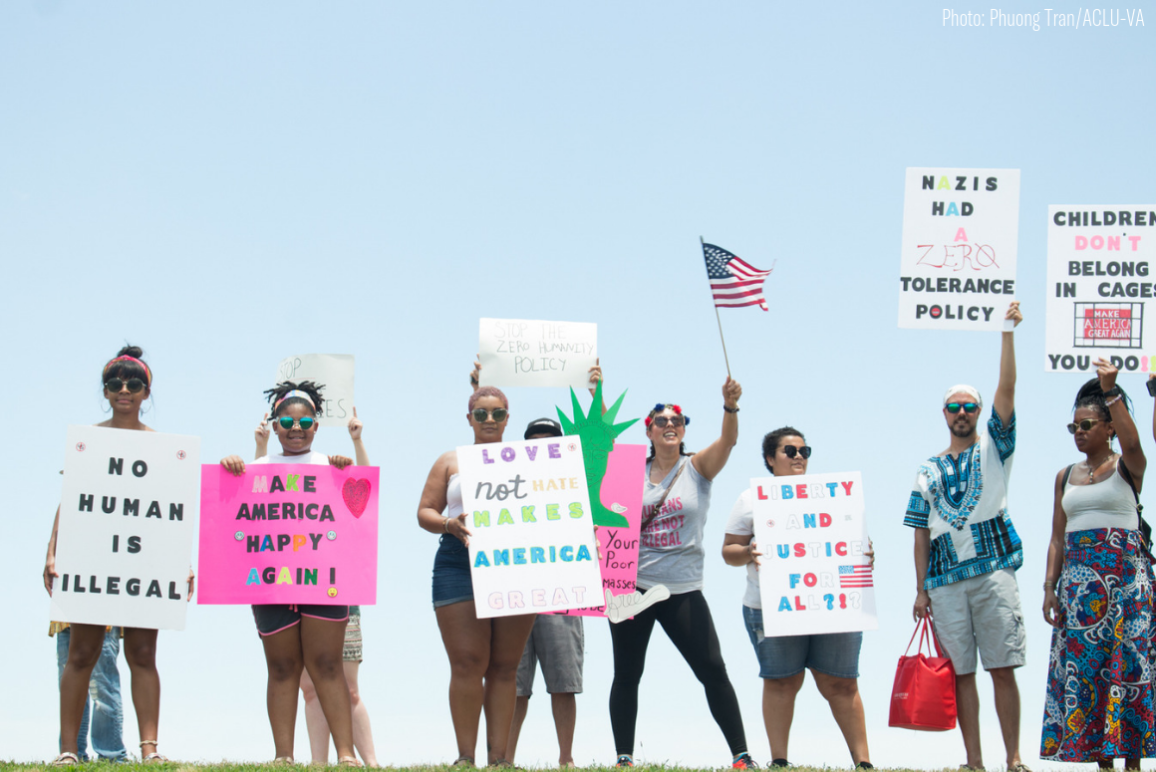A row of protesters holding signs with messages supportive of immigrants ("No Body is Illegal" "Make America Happy Again" "Love Not Hate Makes America Great" etc)