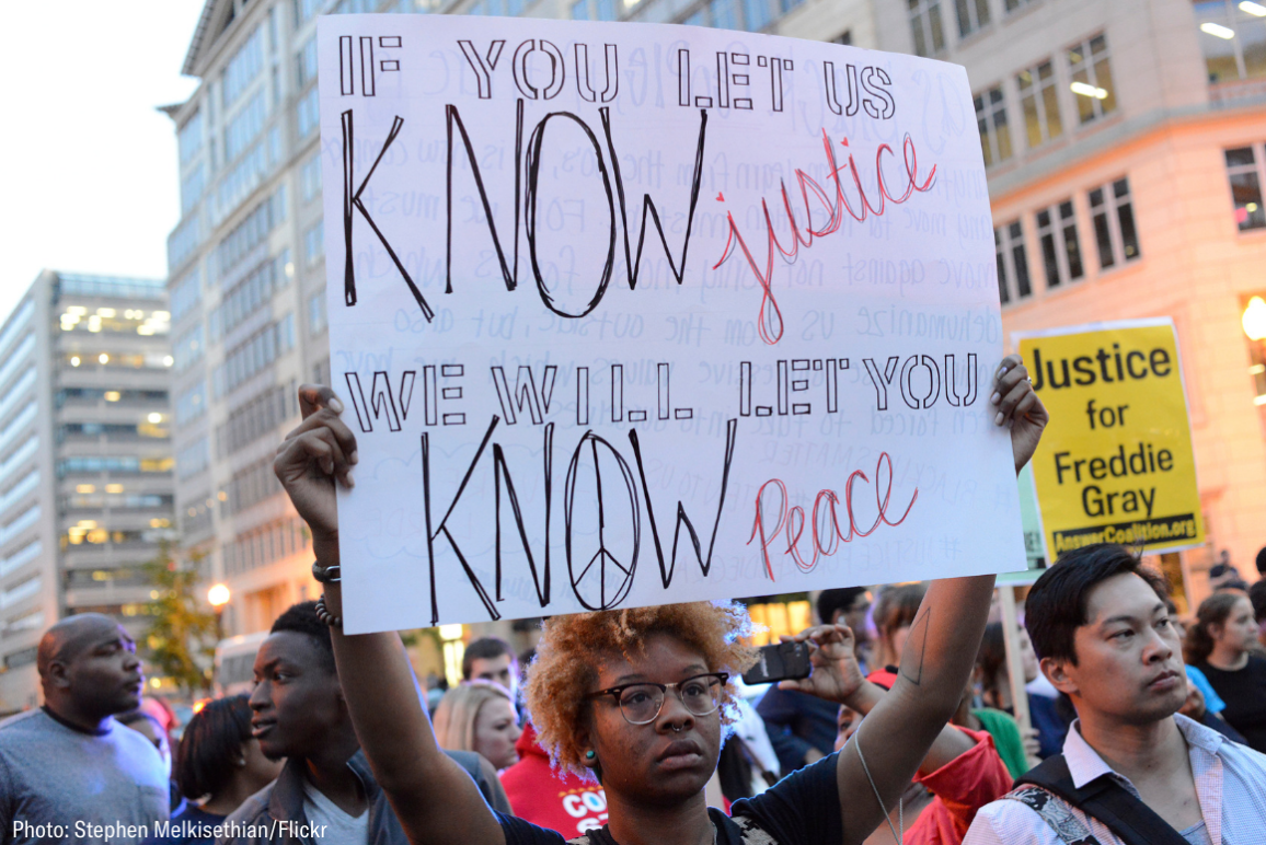 a Black protester holding a sign that says "if you let us know justice, we will let you know peace"