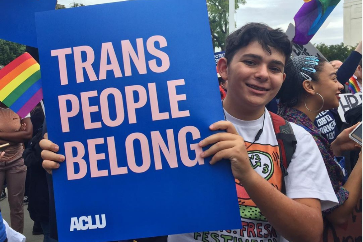 a Hispanic young man holding a sign that says "Trans People Belong"