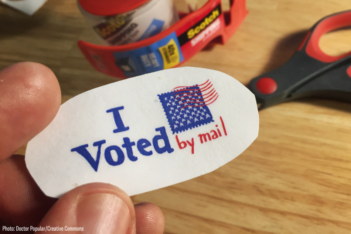 A hand holding a sticker that says "I Voted By Mail"
