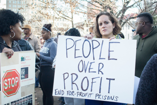 a white woman holding a sign that says "People Over Profit: End For-Profit Prisons)