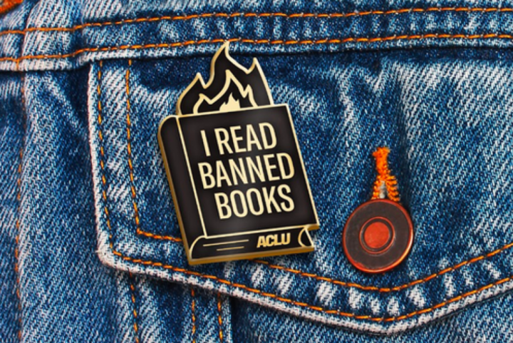 an ACLU branded "I read banned books" pin with fire coming out of a book, against a blue jeans jacket pocket.