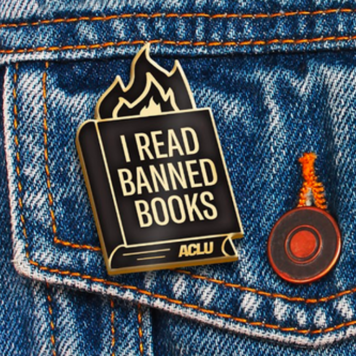 an ACLU branded "I read banned books" pin with fire coming out of a book, against a blue jeans jacket pocket.